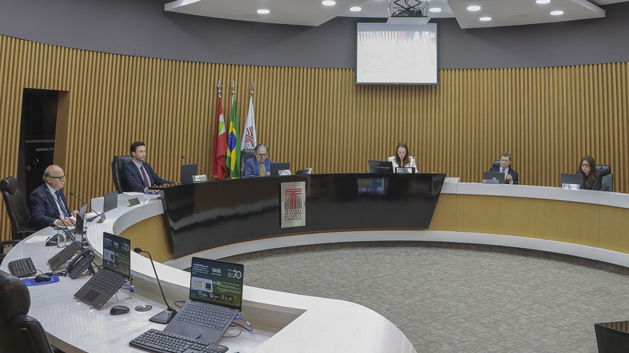 Banner horizontal com foto do Plenário do TCE/SC. Há várias autoridades sentadas diante de bancada circular, com computadores e microfones. Ao centro, o presidente da Primeira Câmara, Aderson Flores. 