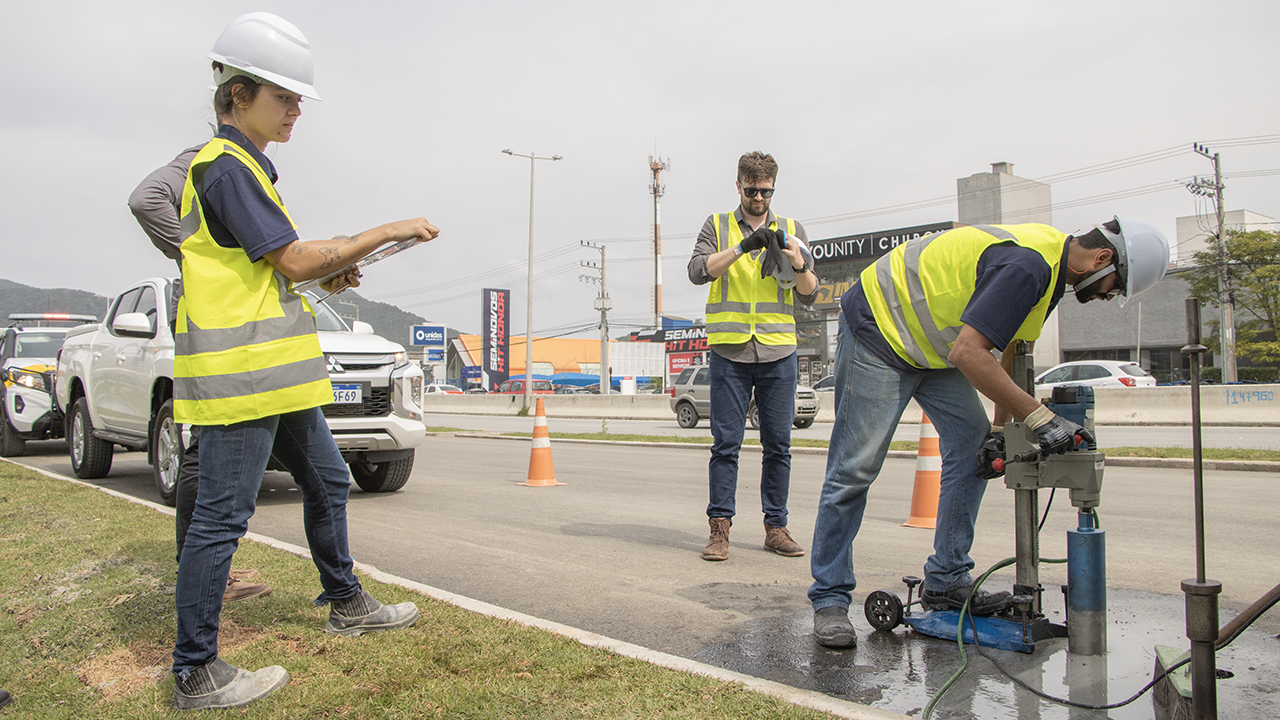 Foto mostra três pessoas em uma rodovia rajando coletes amarelos. A pessoa à esquerda está com uma planilha na mão e de capacete branco. A pessoa ao centro está de óculos escuros e calçando uma luva. A terceira, à direita, também usa capacete e manuseia uma máquina que fura o chão
