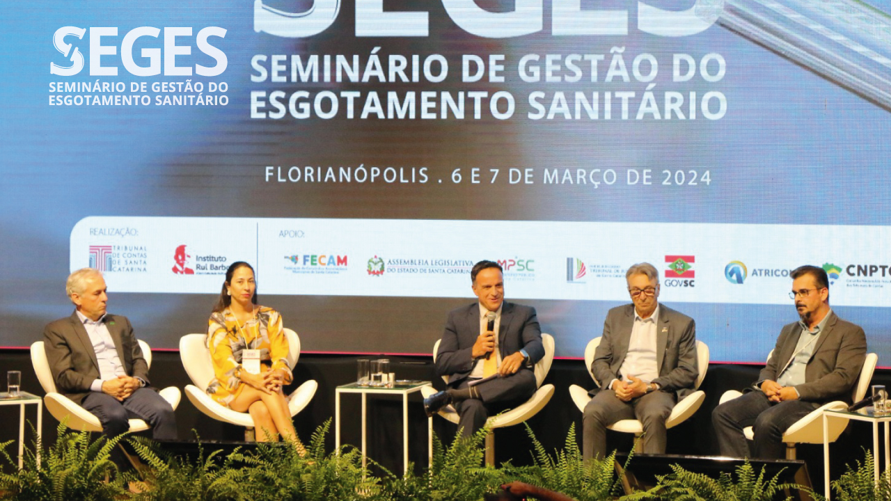 Na foto, os palestrantes estão sentados à frente de uma projeção com o nome do seminário. São quatro homens e uma mulher