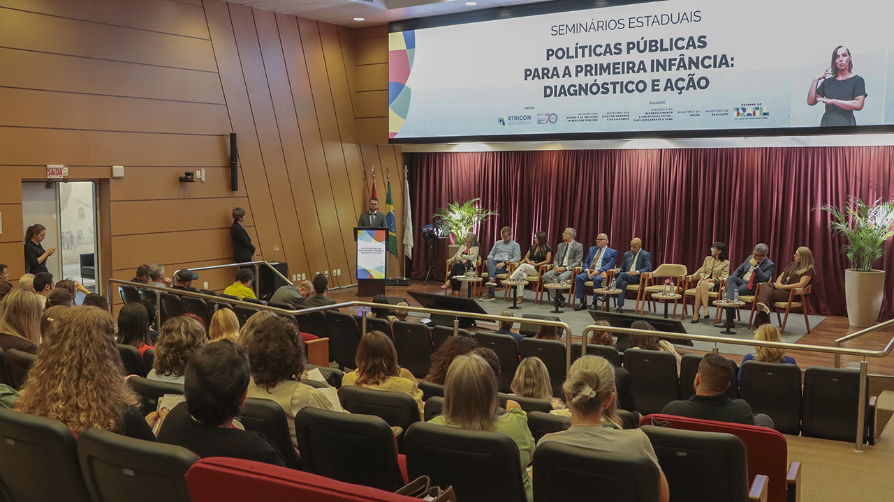Banner horizontal com foto de evento institucional no auditório do TCE/SC. Ao centro, um palco com mesa de autoridades reúne diversos participantes sentados, enquanto um palestrante fala em um púlpito à esquerda. Ao fundo, há uma cortina vermelha e um telão com o título “Seminários Estaduais – Políticas Públicas para a Primeira Infância: Diagnóstico e Ação”. Em primeiro plano, o público ocupa as cadeiras do auditório, assistindo à apresentação. 