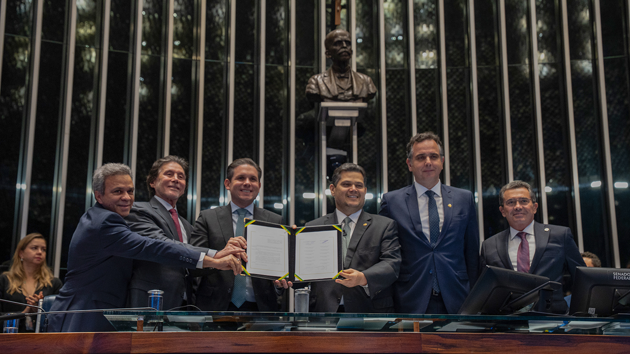 Banner horizontal com foto dos integrantes da mesa de autoridades no Senado segurando um documento. Há seis pessoas, lado a lado, que vestem terno e gravata. Entre os presentes, o presidente do Senado, Davi Alcolumbre, o presidente do TCU, ministro Vital do Rêgo, e o presidente da Atricon, Edilson Silva. 