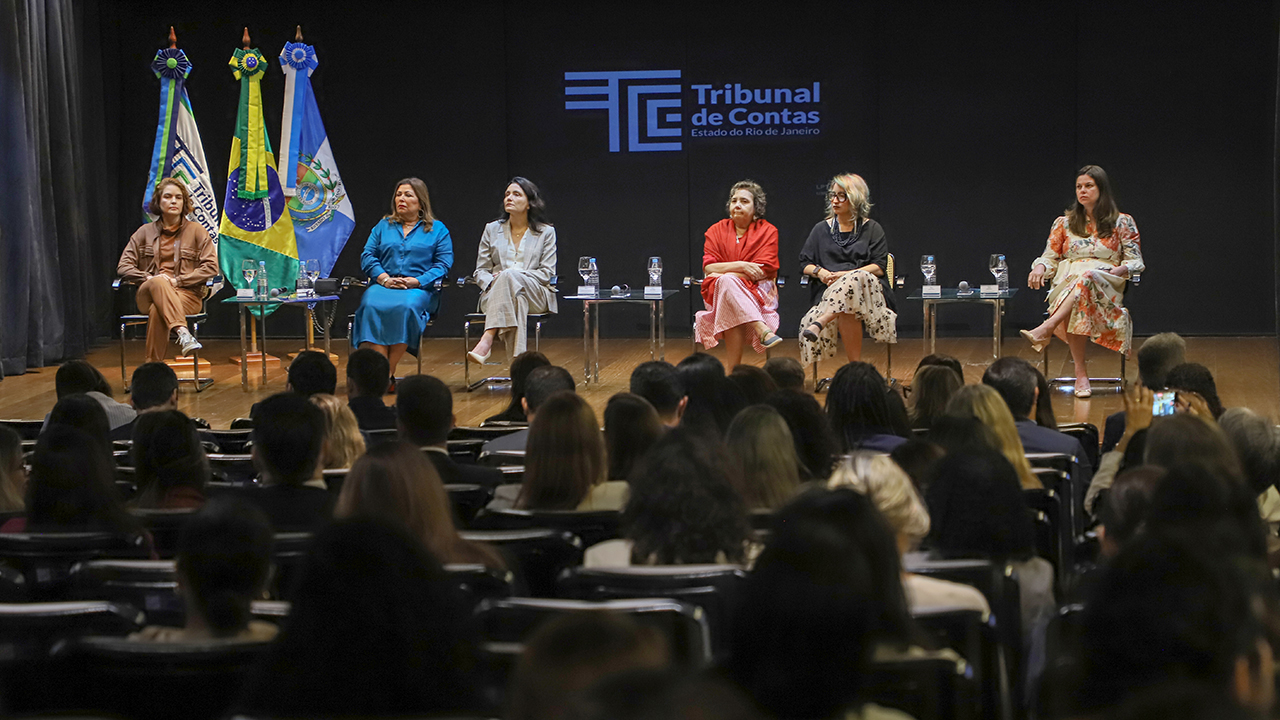 Banner horizontal com foto da plateia acompanhando um painel de debates realizado no TCE-RJ. No palco, seis mulheres estão sentadas em poltronas dispostas lado a lado, com mesas de apoio e copos de água à frente. Entre as palestrantes, está a conselheira substituta do TCE/SC Sabrina Iocken. 