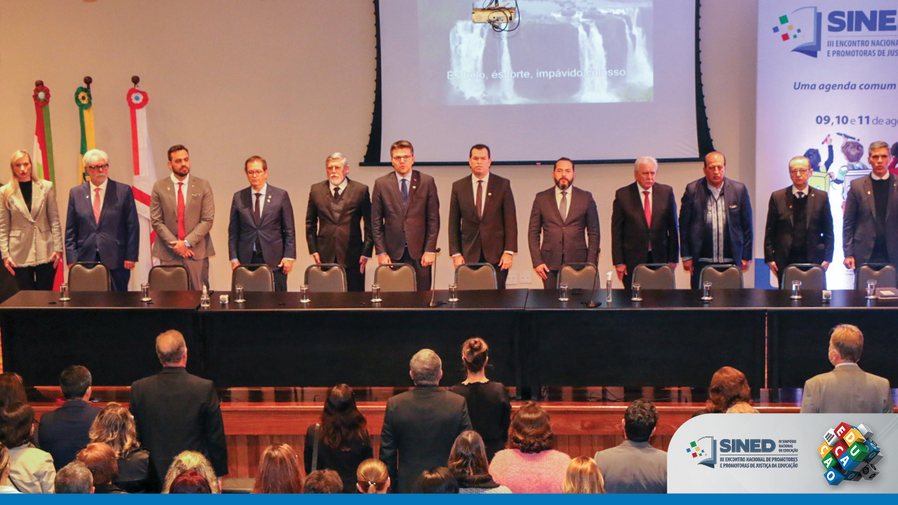 Foto com os 12 integrantes que compuseram a mesa do evento. Ao fundo, à direita, uma parte do painel do Sined, ao centro, a tela de projeção e, à esquerda, as bandeiras nacional, estadual e municipal. À frente dos integrantes da mesa, há parte da plateia. No canto inferior direito da foto, há o logotipo do evento. 