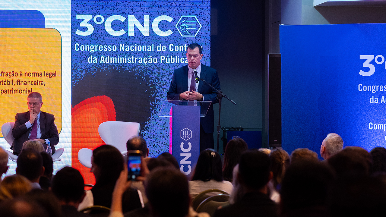 Foto do corregedor-geral do TCE/SC, Adircélio de Moraes, atrás de um púlpito de vidro, palestrando no Congresso. Ele veste terno azul marinho, camisa branca e gravata bordô. Ao fundo, parte de um painel, com a logo do evento. Ao lado esquerdo, sentado numa cadeira, está o ministro do TCU, Antônio Anastasia, que veste terso escuro, camisa azul claro e gravata bordô. A frente, aparecem as cabeças de algumas pessoas que assistem à palestra. 