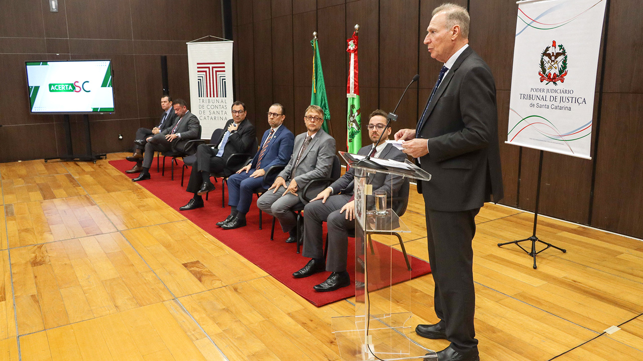 Foto de sete autoridades no palco do auditório, durante o lançamento do Comitê. Há seis homens sentados, entre eles, o conselheiro Adircélio Ferreira Júnior. Em pé, falando ao púlpito, o presidente do TCE/SC, conselheiro Herneus De Nadal. Ao fundo, há banners do TCE/SC e do TJSC.