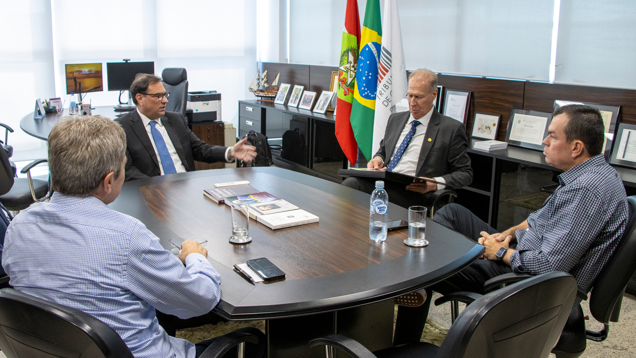 Imagem mostra quatro homens ao redor de uma mesa escura. Dois estão de frente, um de lado e outro de costas. Ao fundo, há um móvel com alguns certificados e fotos e três bandeiras em mastros