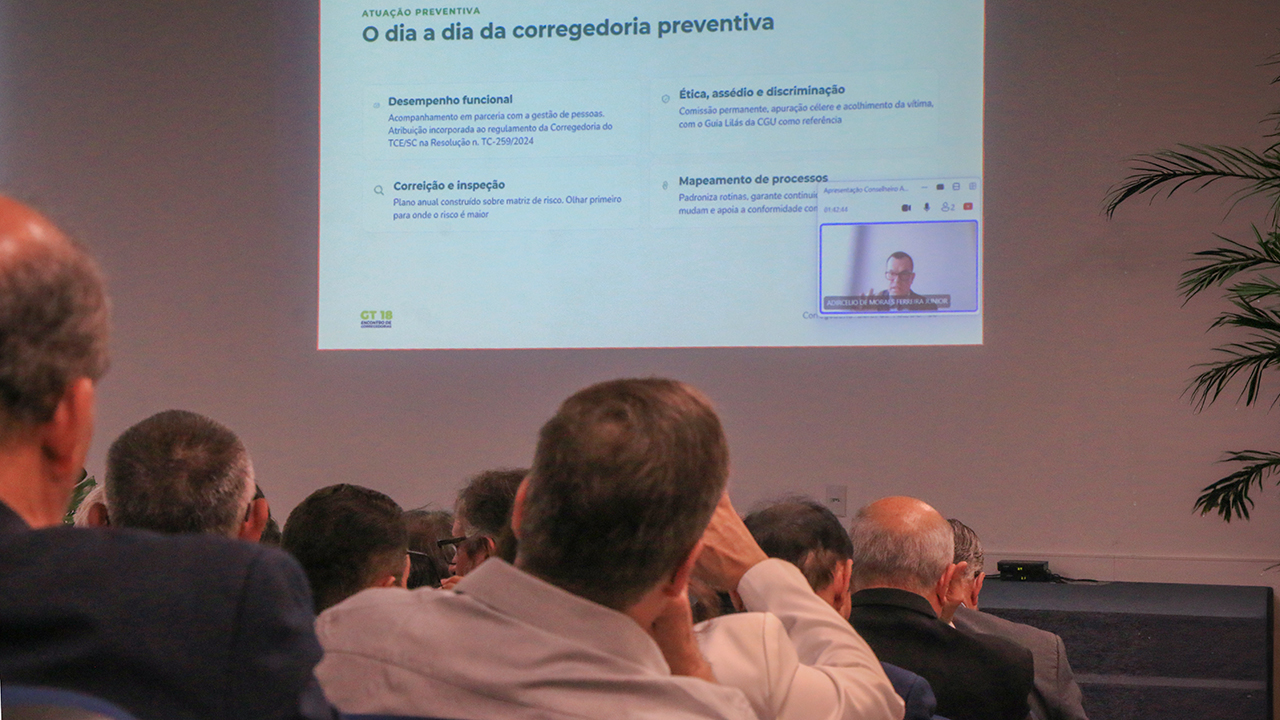 Banner horizontal com foto, em primeiro plano, da plateia assistindo à videoconferência com o conselheiro Adircélio, exibida no telão ao fundo. 