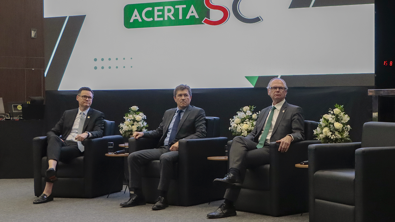 Banner horizontal com do foto conselheiro José Nei Alberton Ascari, ao centro, com o presidente do TJSC, desembargador Rubens Schulz, à direita, e o secretário de Estado da Fazenda, Cleverson Siewert, à esquerda. Eles estão sentados em poltronas no palco do auditório do TJSC. Ao fundo, no telão, é exibido o logo do Acerta SC.