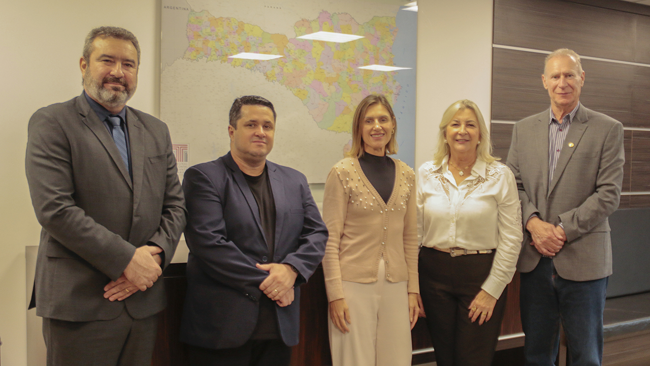 Banner horizontal com foto posada dos integrantes da visita. Em pé, o presidente do TCE/SC, conselheiro Herneus De Nadal, e a diretora de Contas de Gestão, Cláudia Vieira. Ao lado deles, os integrantes da Afamesc, Márcia Zilá e Miquéas Libório. Eles estão em um ambiente institucional. Ao fundo, quadro com mapa de Santa Catarina. 