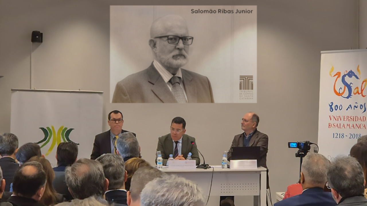 Banner horizontal com foto de três homens sentados diante de uma bancada, no palco. Entre eles, o conselheiro Adircélio Ferreira Júnior e o professor Antonio Arias Rodríguez. À frente deles, o público que assiste ao painel, de costas. Ao fundo, atrás dos painelistas, no telão, foto, em preto e branco, do conselheiro Salomão Ribas Junior. 