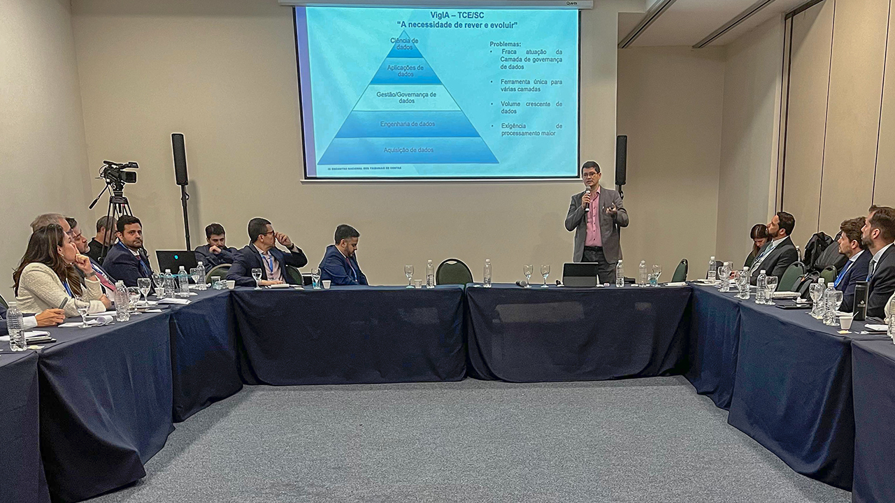 Foto do servidor do TCE/C Alessandro Marinho está fazendo a apresentaação sobre o VigIA.  As cadeiras e mesas estão dipostas em formato de U. Ao fundo do palestrante, um telão exibe a apresentação dele.
