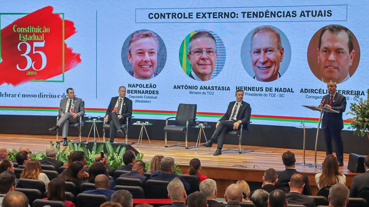 Banner horizontal com foto, em primeiro plano, da plateia que assiste ao painel “controle externo: tendências atuais”. À frente do público, no palco, os conselheiros Adircélio de Moraes e Herneus De Nadal, o deputado Napoleão Bernardes e o ministro do TCU Antônio Anastasia. Atrás deles, é projetado o título do painel e a foto dos participantes. 