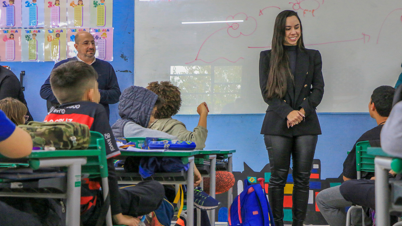 Banner horizontal com foto de uma sala de aula com alunos sentados em suas carteiras durante uma atividade escolar. Em destaque, a auditora Letícia Spindola de Faria está em pé à frente da turma, sorrindo e interagindo com os estudantes. Ao fundo, próximo ao quadro branco, o auditor Renato Bossle Miguel acompanha a atividade. 