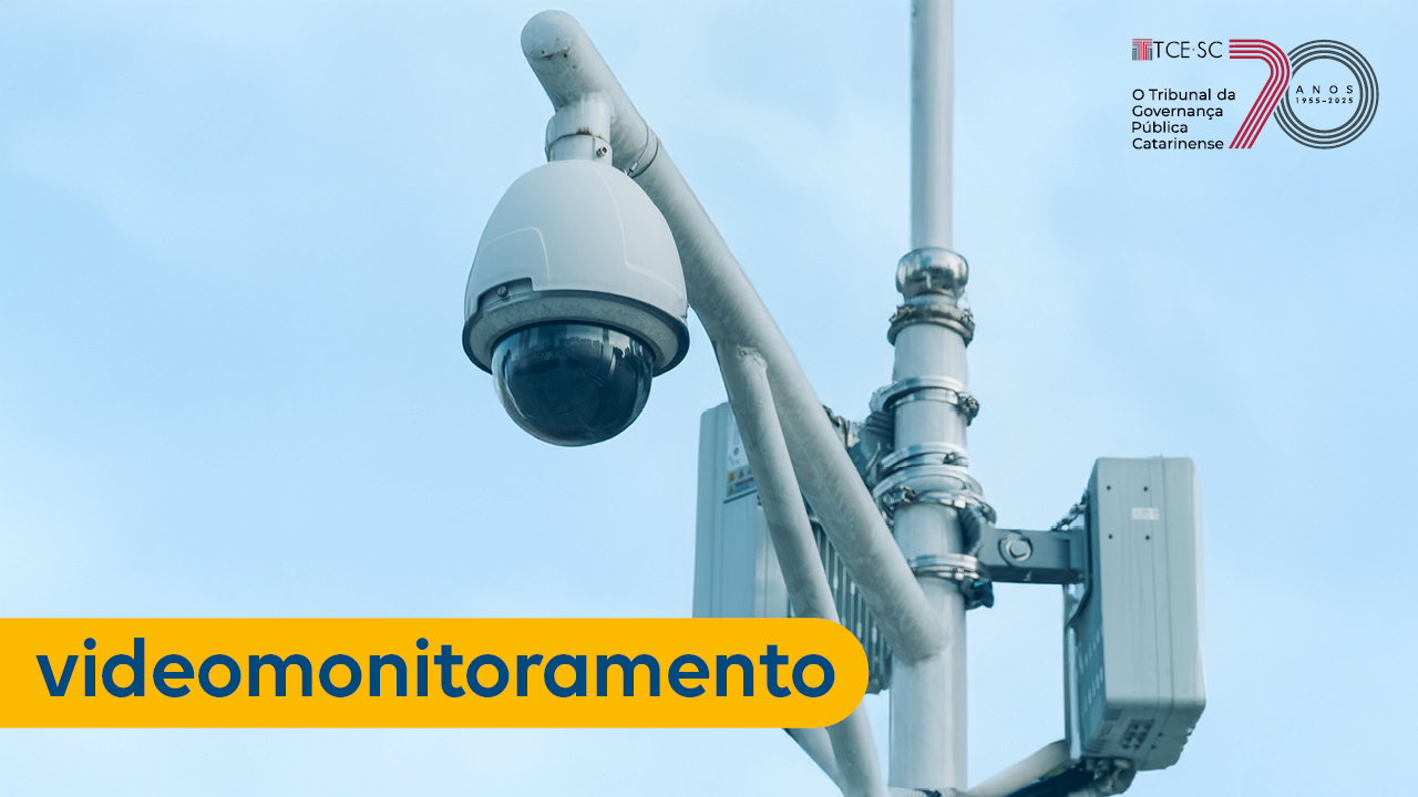 Imagem mostra uma câmera de vigilância em um poste. Na parte inferior esquerda, escrito em azul sobre tarja amarela, está a palavra "videomonitoramento".