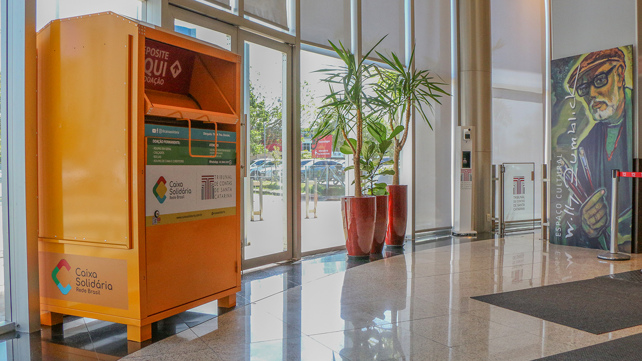 Foto da “caixa solidária” no hall do TCE/SC. A caixa é amarela e contém os logos da Caixa Solidária e do TCE/SC.  