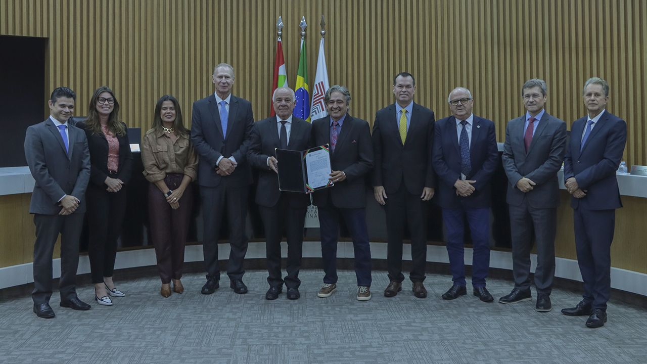 Banner horizontal com foto de conselheiros, conselheiros substitutos e procuradores posicionados lado a lado em ambiente institucional, durante registro formal. Ao centro, estão os presidentes da Primeira e da Segunda Câmara, conselheiros Luiz Eduardo Cherem e Aderson Flores, segurando um documento aberto, em destaque. Ao fundo, há bandeiras oficiais e revestimento em madeira que compõe o plenário.