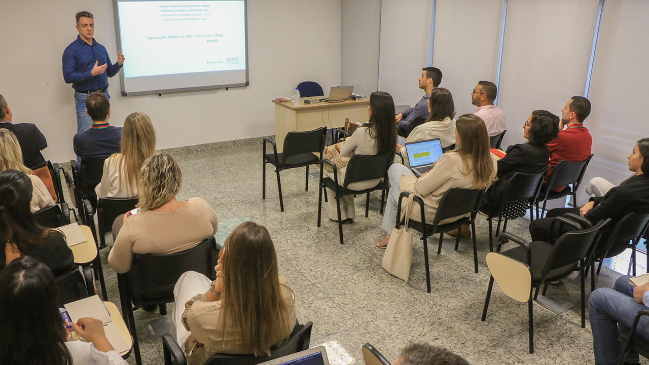 Imagem horizontal que mostra os participantes da Alesc ouvindo a explicação do auditor fiscal do TCE/SC Marcel Belli. Eles estão sentados em carteiras enfileiradas. Marcel está em pé, à frente, próximo ao quadro com projeção da sua apresentação