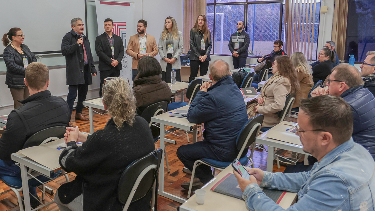 Foto do conselheiro Aderson Flores falando ao microfone durante a etapa de Rio do Sul do Ciclo. Ele é um homem branco, de cabelos claros. Veste um casaco cinza escuro. Ao lado dele, há outras seis pessoas que o acompanham.  Pessoas sentadas nas carteiras assistem apresentação. Há também o banner do TCE/SC. 