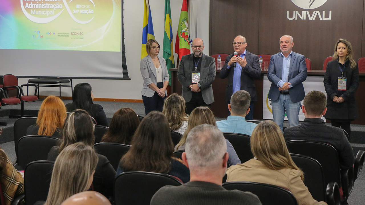Foto da abertura de uma das etapas do Ciclo de Estudos. Em primeiro plano, o público, de costas, que lota o auditório da Univali, sentado em poltronas. À frente, falando ao microfone, membros do TCE/SC. Entre eles, os conselheiros Luiz Eduardo Cherem e Wilson Wan-Dall e o diretor do Icon, Moisés Hoegenn.