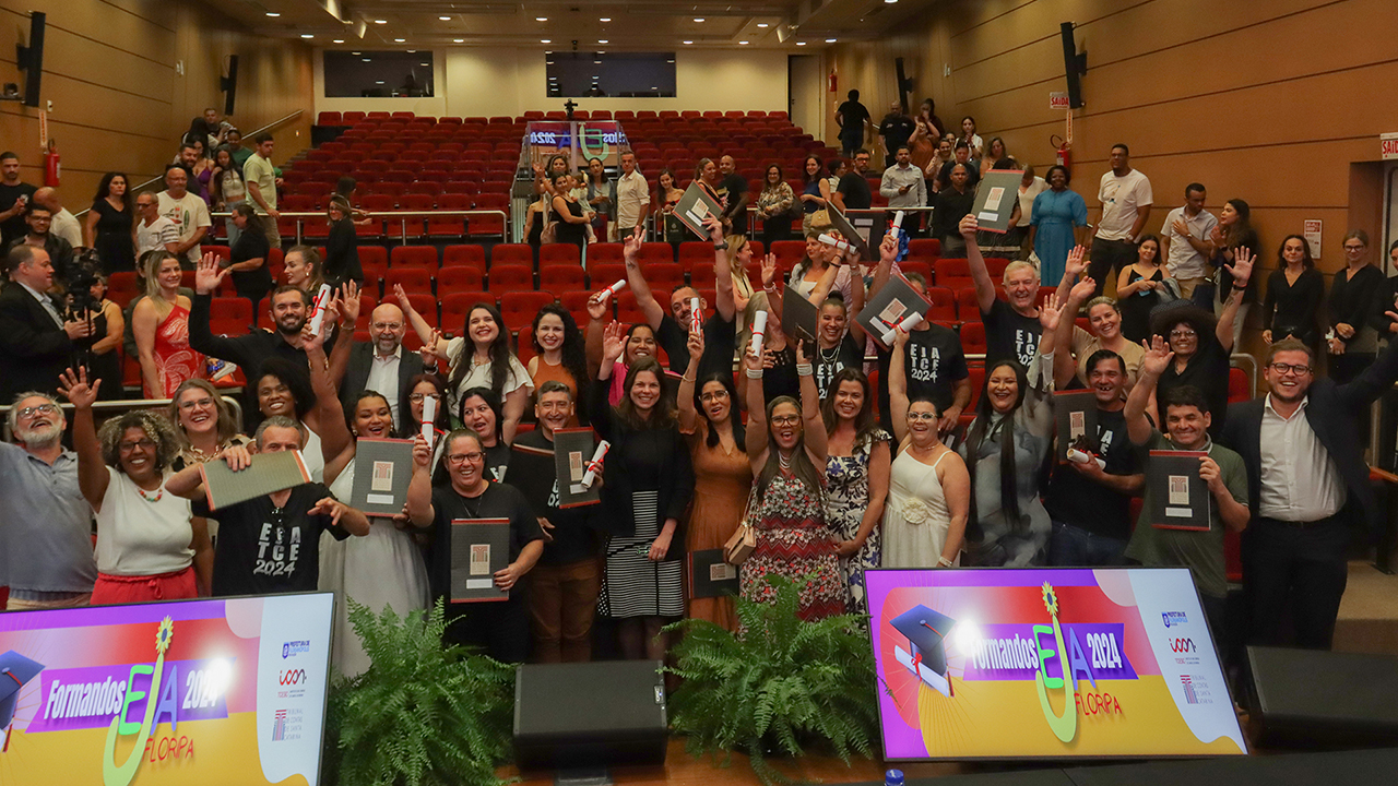 Foto horizontal que mostra os formandos do curso do EJA do TCE/SC em meio à plateia, no auditório do TCE/SC, erguendo os certificados recebidos. Há várias pessoas em pé, em frente às cadeiras vermelhas.  
