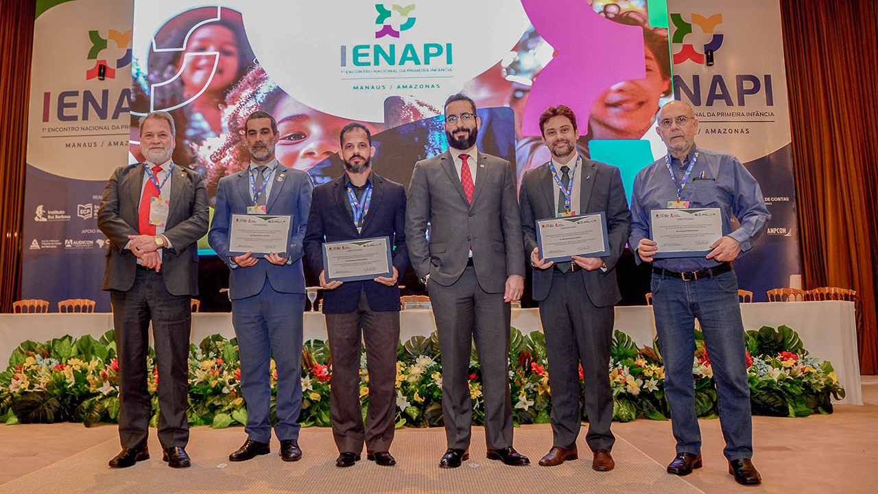 Banner horizontal com foto dos participantes do evento. Há seis homens, em pé, um ao lado do outro, em frente ao banner com logo do primeiro Enapi. Entre os presentes, o fotógrafo Guto Kuerten e os auditores fiscais de controle externo Edelvan da Conceição e Rafael Scherb. Eles seguram certificados.    