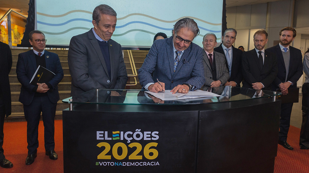 Banner horizontal com foto do conselheiro Aderson Flores assinando o documento, acompanhado por outros representantes ao redor da mesa. Ao fundo, outras autoridades observam a cerimônia. Na parte frontal do púlpito, há a inscrição "Eleições 2026".