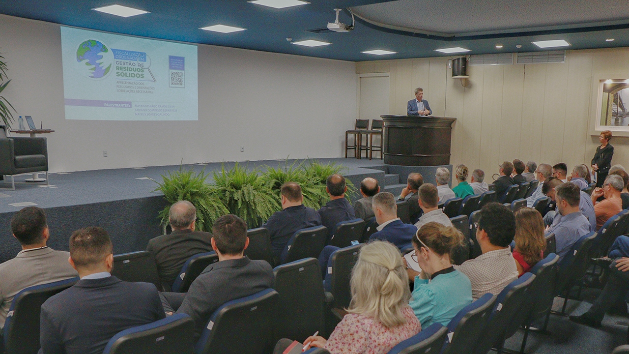 Foto do evento de apresentação de dados relacionados aos resíduos sólidos. Em primeiro plano, de costas, o público sentado em poltronas no auditório azul do TCE/SC. No palco, falando ao microfone na abertura, o conselheiro José Nei Ascari. Ao fundo, a projeção da apresentação. 