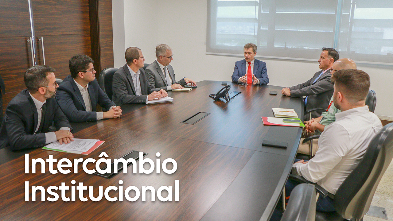 Foto de oito pessoas sentadas ao redor de uma mesa, enquanto conversam, durante a reunião entre o MPSC e o TCE/SC. Todos são homens. Entre eles, estão o conselheiro José Nei Ascari; Paulo Locatelli; e André Milioli. Sobre a mesa, há papéis e copos de água. No canto inferior direito, o título “Intercâmbio Institucional”, em fonte branca. 