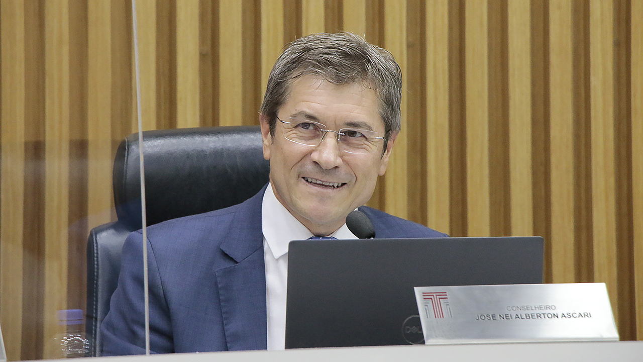 Foto do conselheiro José Nei Ascari no Pleno do TCE/SC. Ele é um homem branco, de cabelos grisalhos. Está vestindo terno azul-escuro, camisa branca e gravata. Usa óculos e está sorrindo ao falar ao microfone. À sua frente, há uma placa de metal com o seu nome e o logo do TCE/SC. 