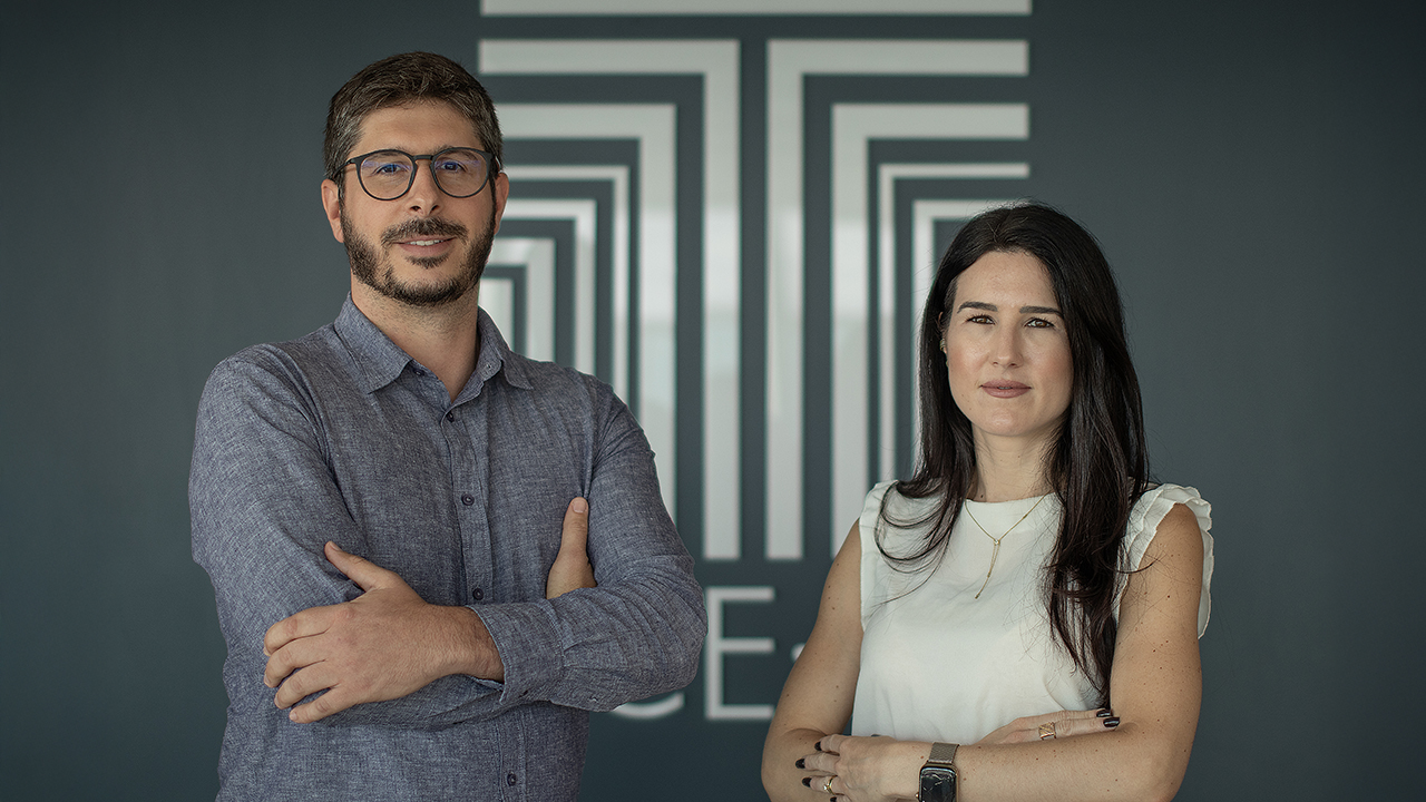 Banner horizontal com foto que mostra os auditores fiscais de controle externo Leandro Marques e Juliana Medeiros das Neves Moser, um ao lado do outro, em pé, com os braços cruzados. Leandro é um homem branco, de cabelos e barba escuros. Veste uma camisa azul e usa óculos de grau. Juliana é uma mulher branca, de cabelos compridos e castanhos. Veste roupa branca. Atrás deles, há uma parede cinza com o logo do TCE/SC na cor branca. 