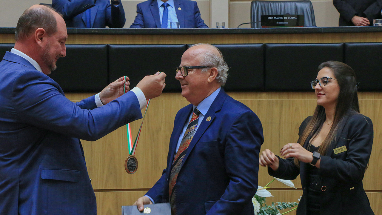 Foto do conselheiro do TCE/SC Wilson Wan-Dall recebe do presidente da Alesc, deputado Mauro de Nadal, a medalha de homenagem à participação na Comstituição Estadual. São os dois homens brancos, que vestem terno azul. Ao lado, uma mulher de cabelos compridos e castanhos auxilia.