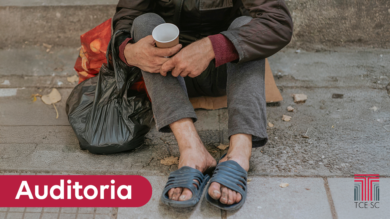 Foto de uma pessoa em situação de rua sentada sobre uma calçada. A foto está cortada, de modo que só é possível ver seus braços e pernas. Ela segura um copo descartável e sacos pretos. Sobre a imagem, no canto inferior esquerdo, o texto “Auditoria”, em tarja vermelha. No canto inferior direito, logo do TCE/SC. 