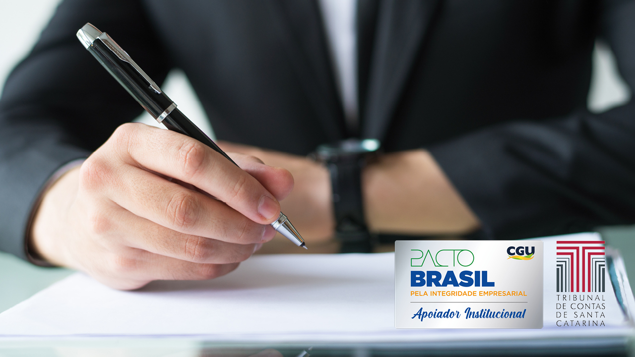 Imagem mostra um homem de casado preto, ao fundo, desfocado, com uma caneta na mão assinando um documento. À direita, no canto inferior, há duas logos. Uma do Pacto Brasil e uma do Tribunal de Contas de Santa Catarina. A do Pacto Brasil tem fundo branco, a palavra “pacto” escrita em verde, letra fina, e a palavra “Brasil” escrita em azul escuro, letras maiores. O logo do TCE é a letra “T” em várias camadas, na cor bordô.