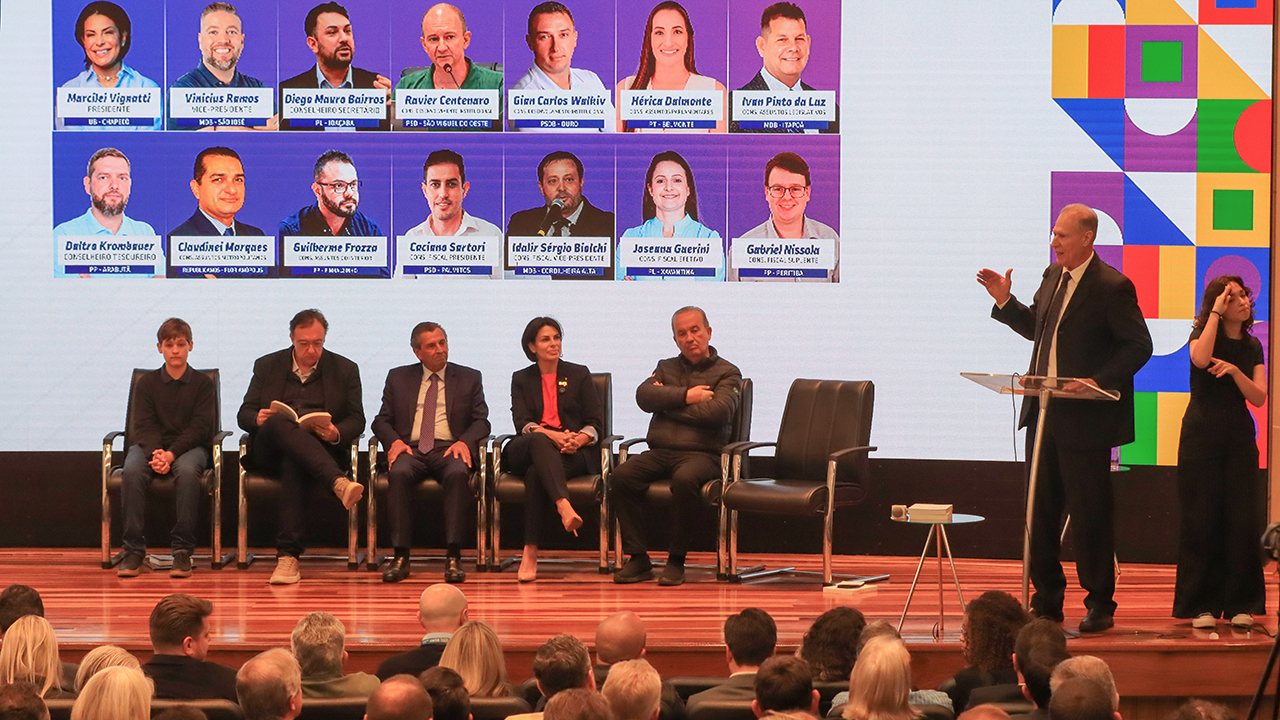 Foto do palco do auditório da Alesc. O presidente do TCE/SC, conselheiro Herneus De Nadal, está no púlpito, ao lado direito da foto, fazendo sua manifestação na posse da diretoria da Uvesc. No palco, estão sentados, da esquerda para a direita, o vereador mirim, João Pedro Probst, a nova presidente da Uvesc,  Marcilei Vignatti; Gilson Conzatti, presidente da UVB; o presidente da Alesc, Julio Garcia,; a presidente da UVESC, Marcilei Vignatti; e o governador Jorginho Mello.