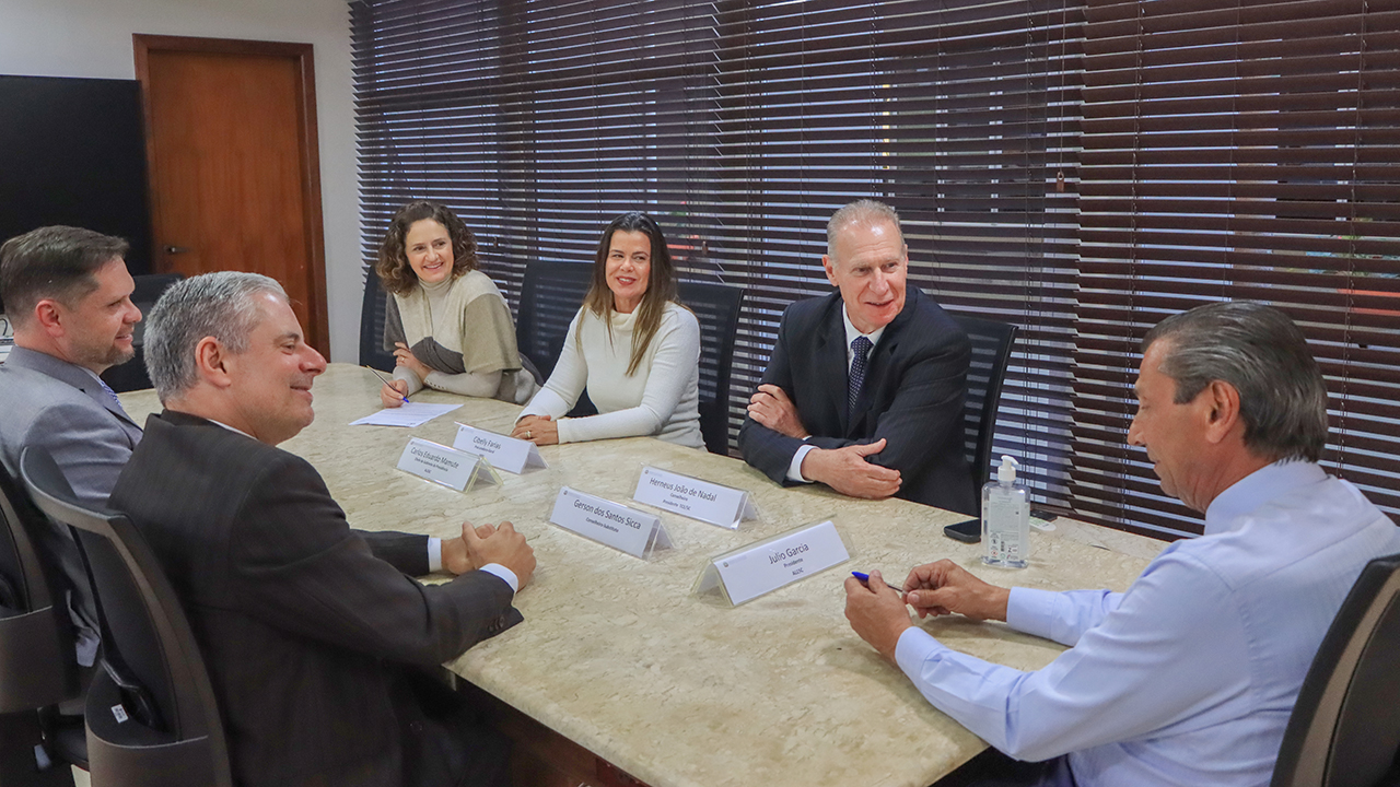 Banner horizontal com foto de seis autoridades reunidas diante de uma mesa enquanto conversam. Entre os presentes, o conselheiro Herneus De Nadal, o conselheiro substituto Gerson Sicca e a procuradora-geral do MPTC/SC, Cibelly Farias. 