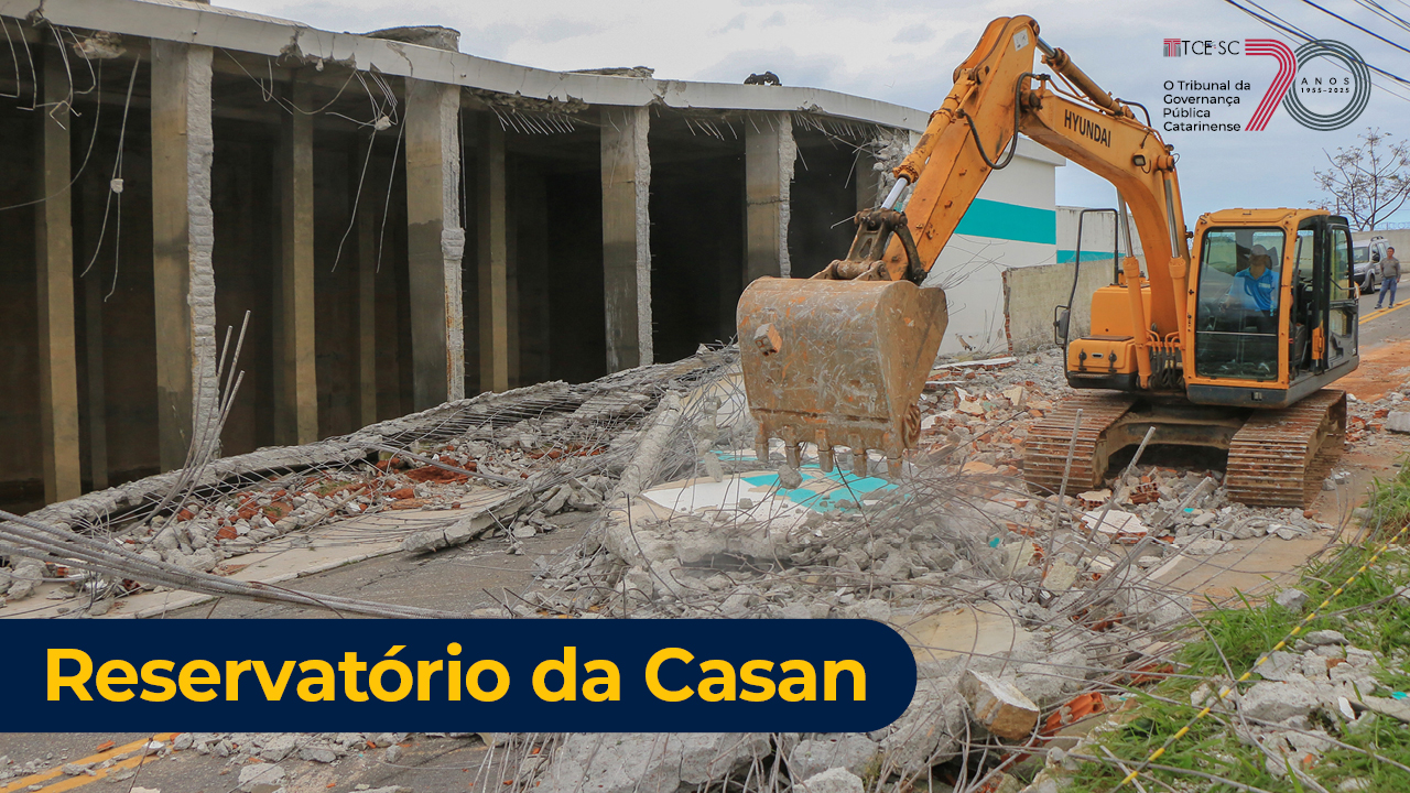 Imagem mostra o local do acidente, com uma estrutura de paredes caídas à esquerda e uma retroescavadeira amarela à direita. No canto inferior esquerta está escrito Reservatório da Casan em letras brancas sobre fundo preto.