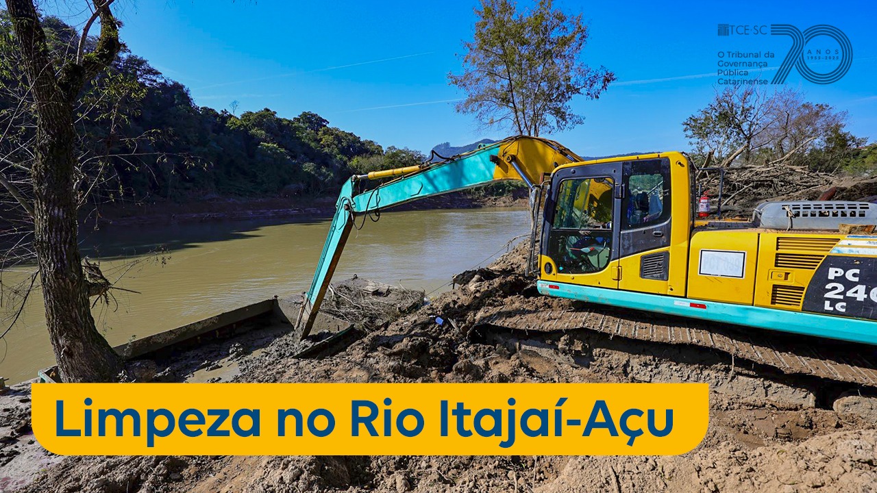 Imagem mostra uma retroescavadeira amarela e azul retirando lama do leito de um rio. Na parte inferior da imagem, está escrito em azul sobre tarja amarela “limpeza no Rio Itajaí-Açu”