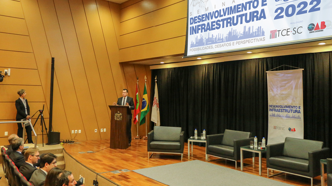 Presidente Adicélio de Moraes Ferreira Júnior faz a abertura do Seminário Desenvolvimento e Infraestrutura. Ele está no púlpito, a esquerda da imagem. No lado direto, há três poltronas, ao fundo, o banner de identificação do evento e, à frente, parte da plateia.