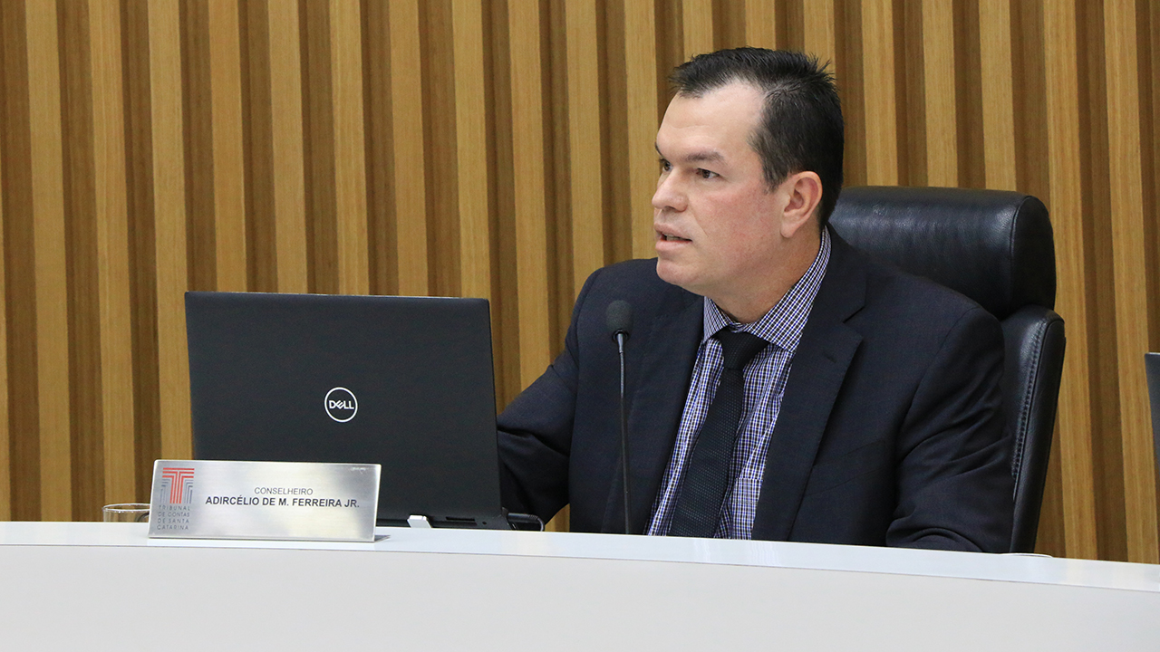 Foto do conselheiro do TCE/SC Adircélio de Moraes Ferreira Júnior, no plenário do TRibunal. Ele é um homem branco, de cabelo escuro, veste um terno preto e usa camisa cinza e gravata escura.