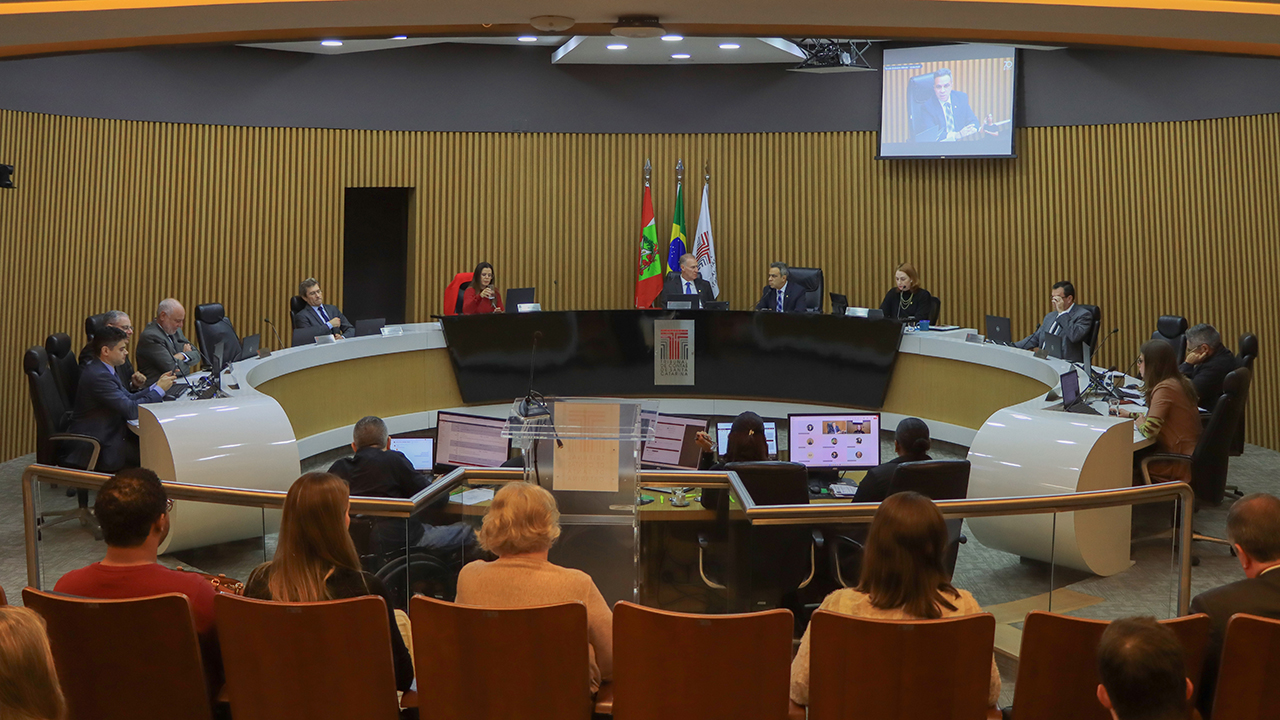 Imagem mostra o plenário do TCE/SC. As paredes são amareladas. Em frente, há uma mesa em forma de U onde ficam os conselheiros. Há televisões nas paredes onde ficam projetadas as imagens da própria sessão.