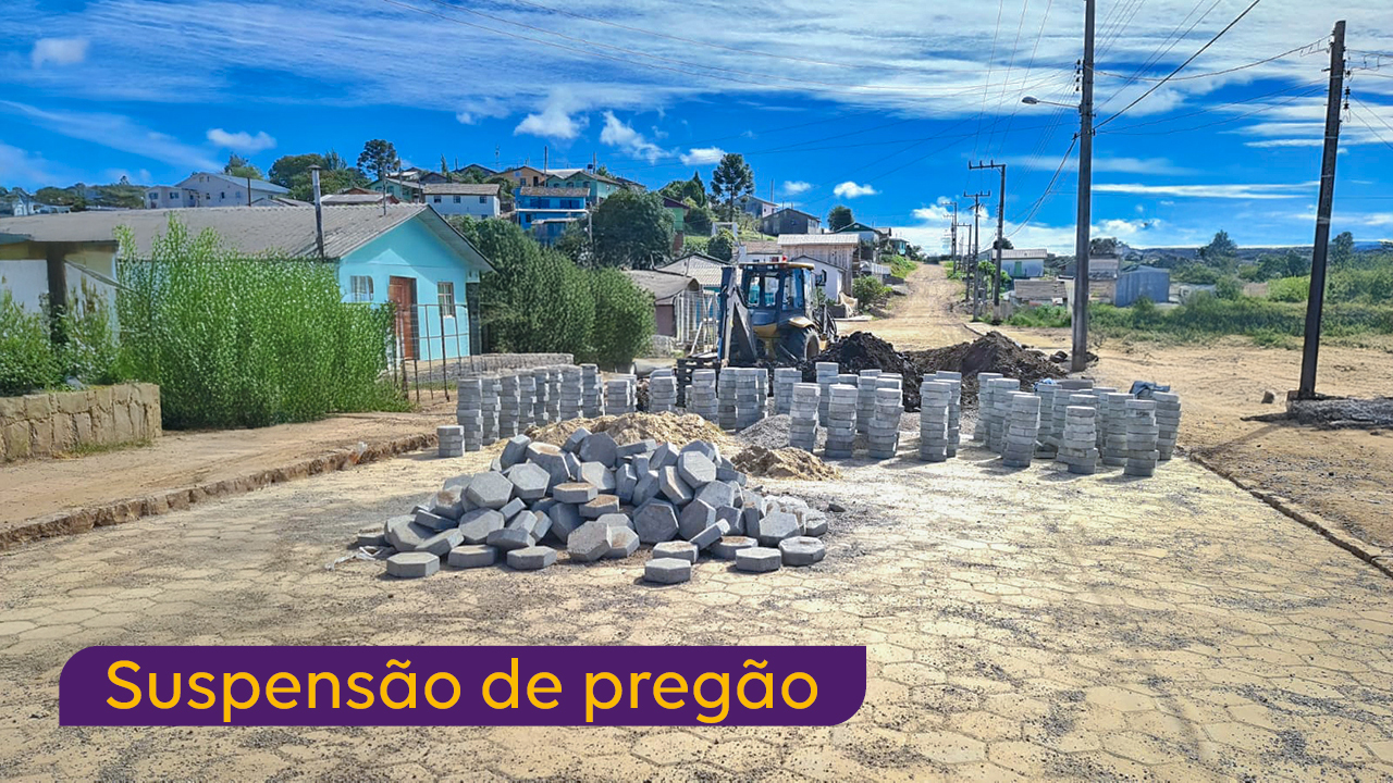 Foto de uma rua, vista em perspectiva, sendo calçada com lajotas. Na metade anterior da rua, o serviço já foi executado. No meio, há pilhas de lajotas, e do meio para trás, a rua é de barro e há uma retroescavadeira trabalhando. No lado direito da foto, algumas casas, e no lado esquerdo, postes. Sobre a foto, no acanto inferior esquerdo, uma tarja cor de vinho com o título “Suspensão de pregão”. 