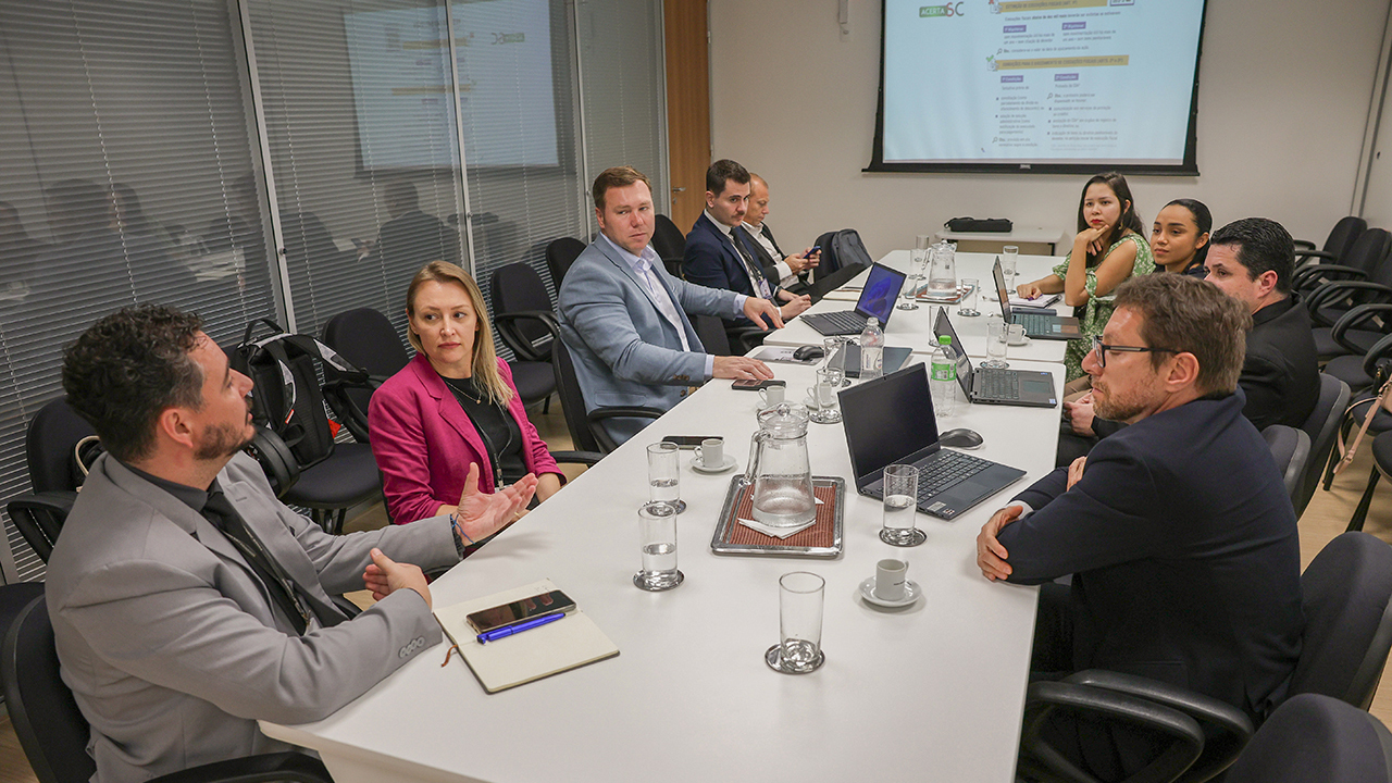 Imagem é de uma reunião com nove pessoas ao redor de uma mesa branca. São seis homens e três mulheres. Sobre a mesas há laptops e copos de água. Ao fundo há um telão