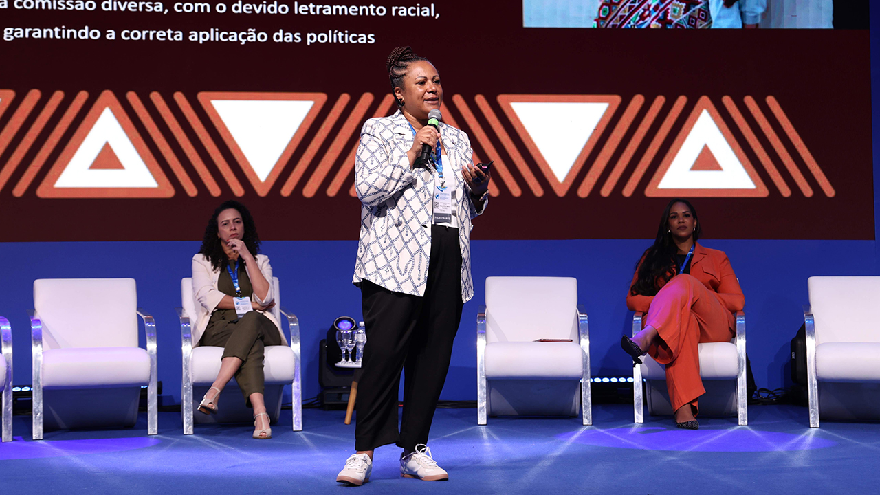 Foto da diretora Walkíria Machado Rodrigues Maciel proferindo palestra no evento. Ela é uma mulher negra. Está em pé, no palco do evento, falando ao microfone. Atrás dela, outras participantes, sentadas em poltronas. No telão, ao fundo, é projetada a apresentação.