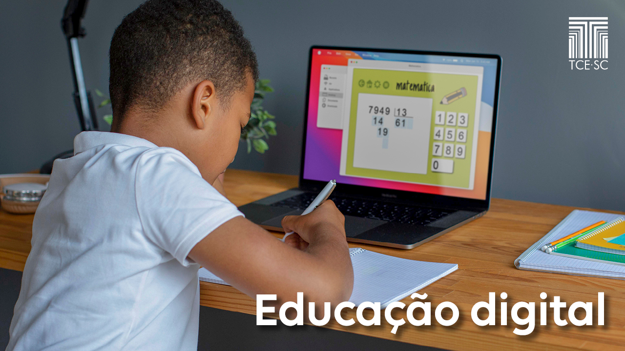 Imagem mostra uma criança de costas, vestindo camiseta branca e em frente a um computador que está aberto sobre uma mesa. A criança segura um lápis e escreve em um caderno. O computador mostra uma conta de matemática em sua tela.