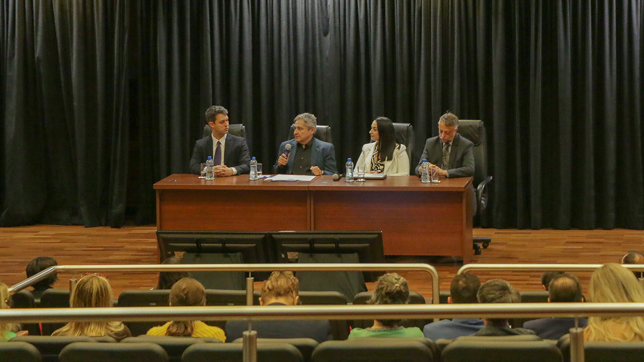 Foto da abertura do 1.° Congresso Nacional "Desafios de Implementação do ESG". Estão sentados diante de uma bancada, um ao lado do outro, no palco do auditório do TCE/SC, da esquerda para a direita, Fabiano Sponholz Araujo, presidente do Ceprev, o conselheiro Aderson Flores, do TCE/SC, Gabriela Giacomolli, coordenadora científica do Congresso, e Diogo Ringenberg, procurador-geral do MPTC/SC. O conselheiro Aderson fala ao microfone. À frente do palco, de costas para a foto, há pessoas que assistem à abertura