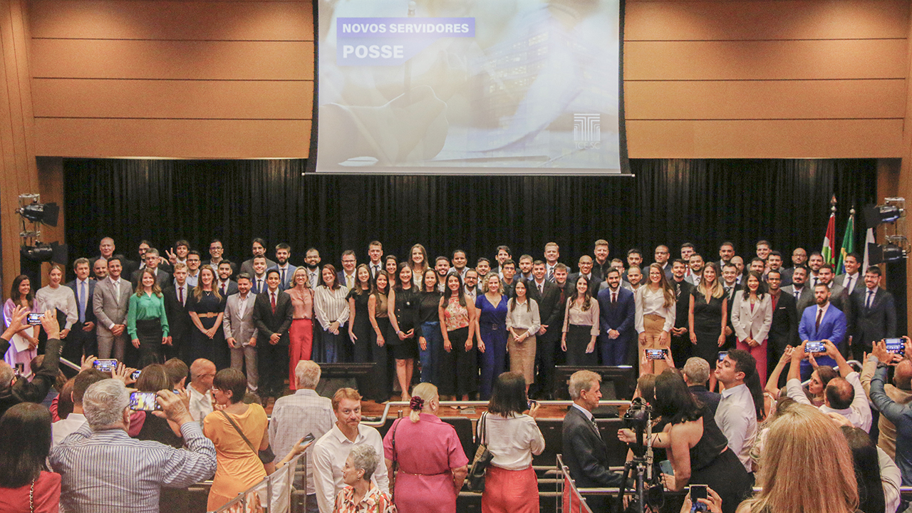 Os 68 novos servidores (mulheres e homens), em pé, no palco do auditório do TCE/SC. Ao fundo e no alto, telão, com a imagem do banner que identifica o concurso. A frente, algumas pessoas, da plateia, em pé. 