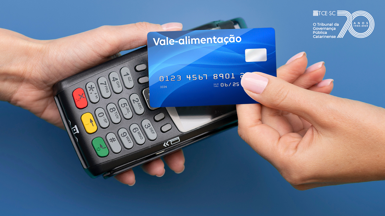 Imagem em fundo azul mostra uma mão segurando uma máquina de cobrança e outra mão segurando um cartão de pagamento. Nesse cartão está escrito Vale-alimentação. No alto, à direita, já o logo alusivo aos 70 anos do TCE/SC 