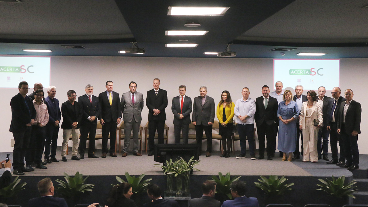Foto das autoridades no palco do auditório do TCE/SC, durante reunião do Comitê Gestor do Acerta SC. Há 21 pessoas em pé, uma ao lado da outra. Entre os presentes, os presidentes Herneus De Nadal e Francisco Oliveira Neto, os conselheiros Aderson Flores, Adircélio de Moraes e José Nei Ascari e a procuradora-geral adjunta Cibelly Farias.