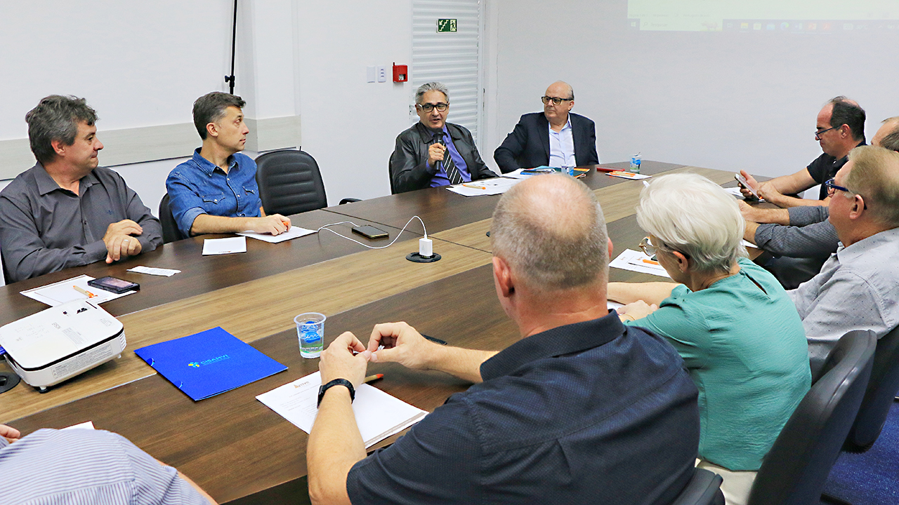 Imagem mostra várias pessoas ao redor de uma mesa. Ao fundo, falando ao microfone, o conselheiro Aderson Flores. Ao lado direito dele está o conselheiro Wilson Wan-Dall