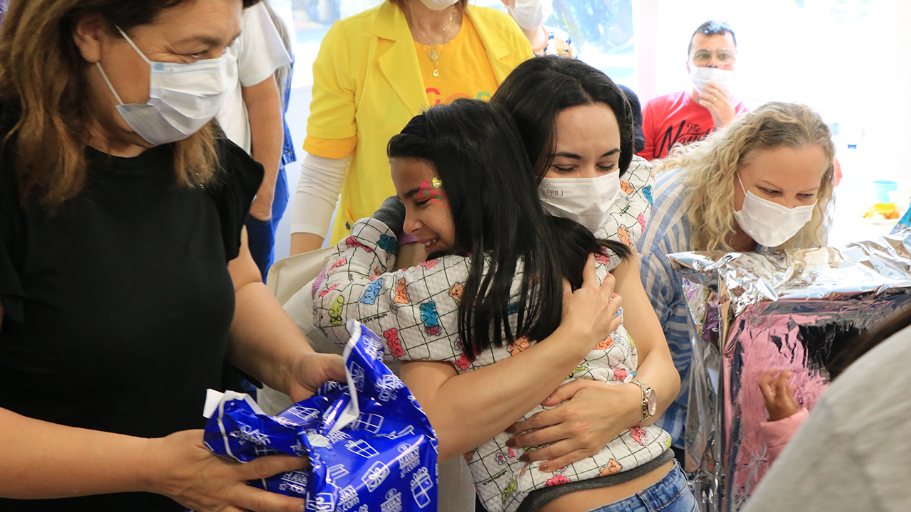 Foto de três servidoras do TCE/SC entregando presentes às crianças que estão em tratamento oncológico no Hospital Infantil Joana de Gusmão. À esquerda, há uma mulher segurando um pacote de presente azul e, à direita, uma mulher segurando um pacote de presente prata. Ao meio, uma mulher está abaixada, abraçando uma menina, que sorri. Ao fundo, há mais pessoas.